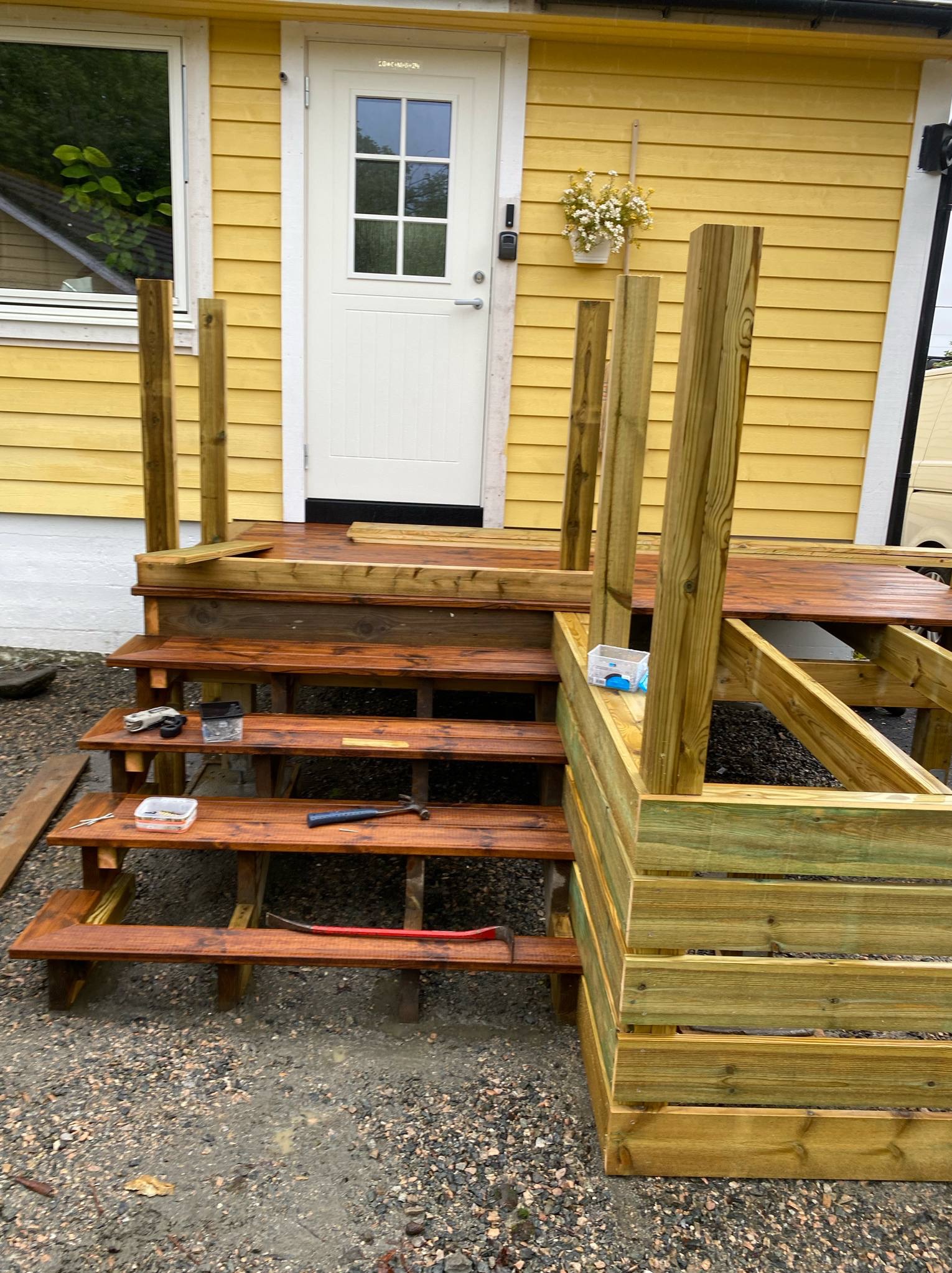 Budowa drewnianego tarasu z schodami przy żółtym domu. Widoczne narzędzia i elementy konstrukcyjne w trakcie montażu. Częściowo zadaszony taras w trakcie budowy.