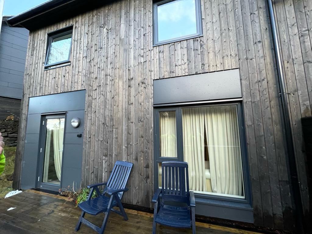 Nowoczesny dom szkieletowy z drewnianą elewacją i tarasem. Dwa niebieskie krzesła na tarasie. Okna z białymi zasłonami. Ciemna kolorystyka.