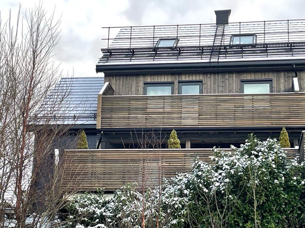 Dwukondygnacyjny dom z drewnianą elewacją i balkonem z poziomymi deskami, częściowo pokryty śniegiem. Widoczne krzewy i drzewa bez liści.