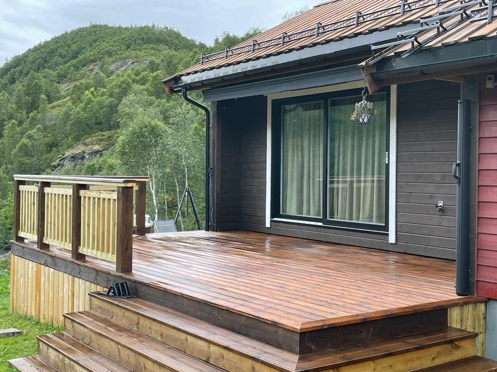 Nowoczesny taras drewniany z balustradą, przylegający do domu z przesuwnymi drzwiami. Widok na górzysty krajobraz w tle. Drewno po impregnacji, widoczne krople wody.