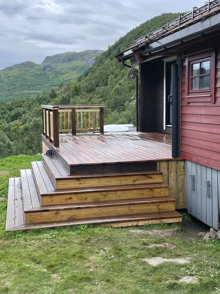 Drewniany taras z balustradą i schodami przy czerwonym domu na tle górskiego krajobrazu. Widoczne detale wykończenia i naturalne otoczenie.