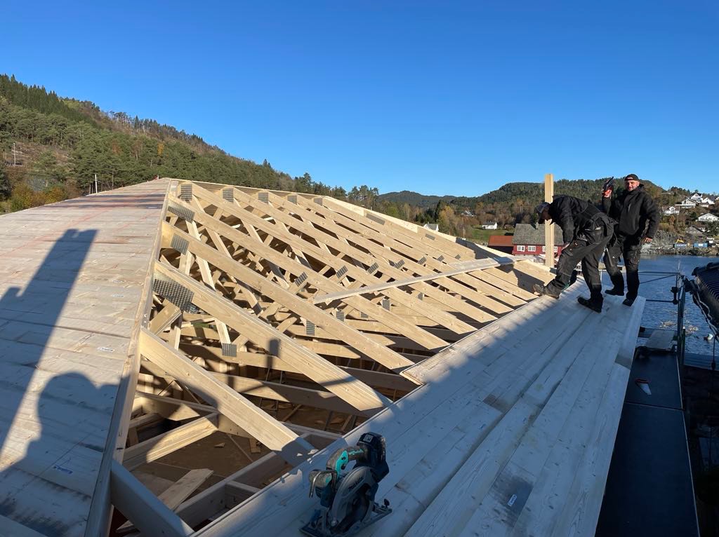 Konstrukcja drewniana dachu w trakcie budowy, widoczni pracownicy i narzędzia, malownicze tło z zielenią i wodą. Ujęcie z góry, w słoneczny dzień.