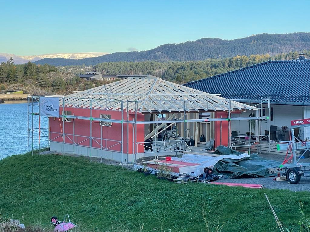 Budowa domu szkieletowego w malowniczej scenerii: konstrukcja z czerwonymi panelami, drewniany szkielet dachu, rusztowania i przyczepa na tle jeziora i gór.