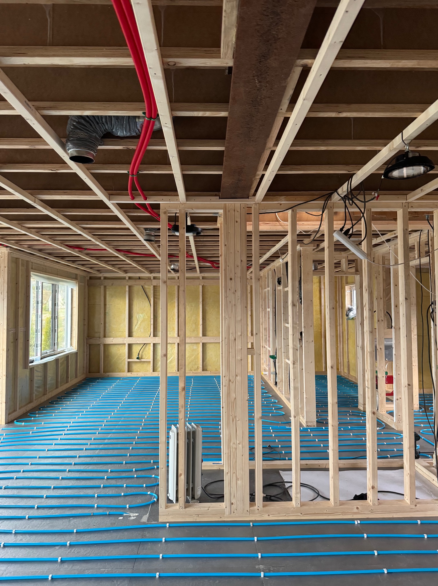 Wnętrze domu w trakcie budowy: drewniany szkielet, ocieplenie ścian wełną mineralną, instalacja ogrzewania podłogowego z niebieskich rur. Widoczne okno i elementy wentylacji.