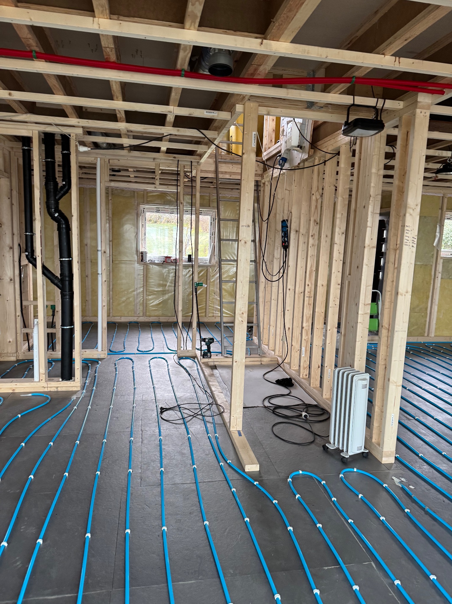 Wnętrze domu w trakcie budowy: drewniana konstrukcja szkieletowa, instalacja ogrzewania podłogowego z niebieskich rur, widoczne elementy wentylacji i instalacji elektrycznej.