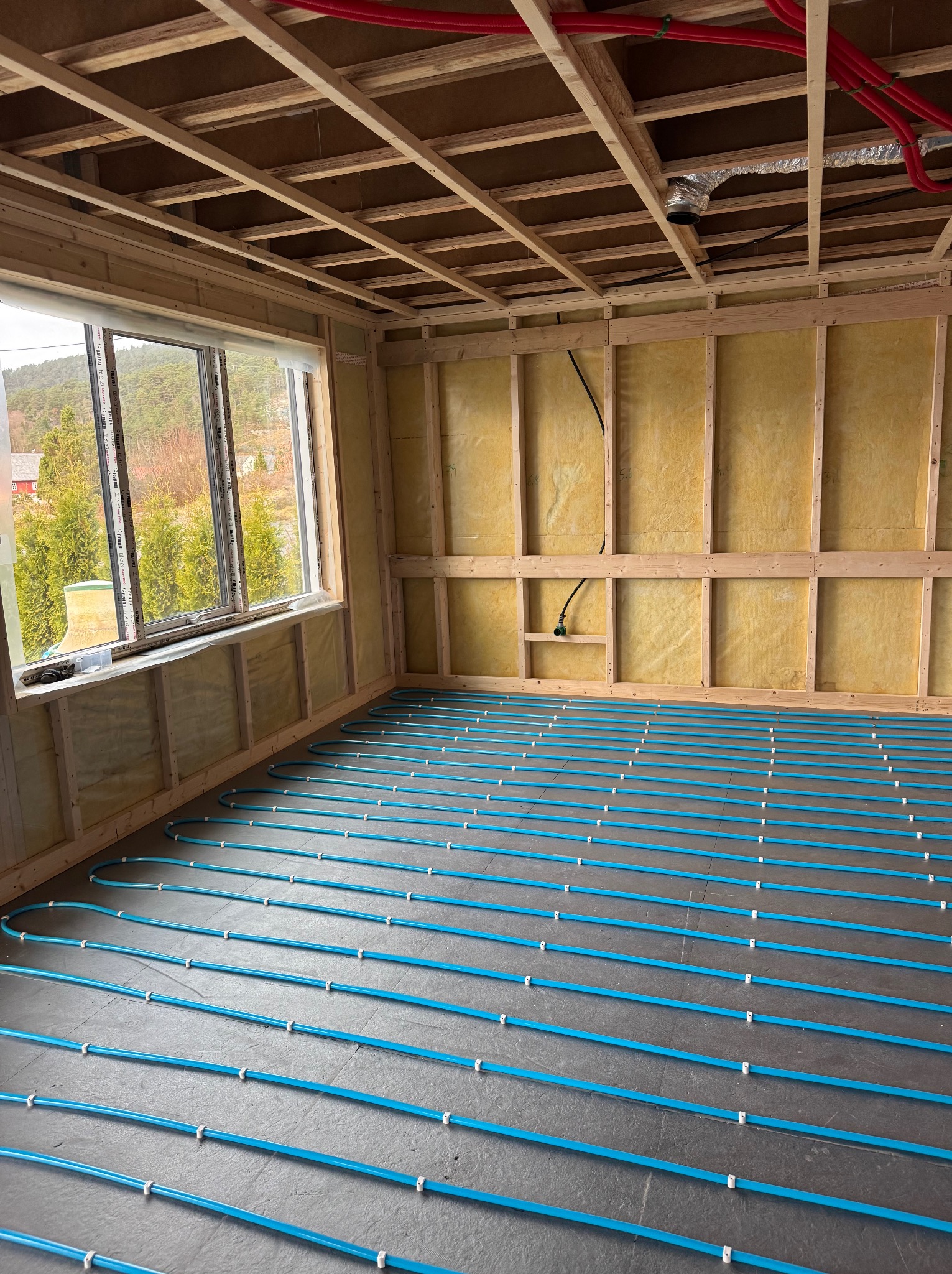 Wnętrze domu szkieletowego w trakcie budowy: izolowane ściany, drewniana konstrukcja stropu i niebieskie rury ogrzewania podłogowego na szarej posadzce.