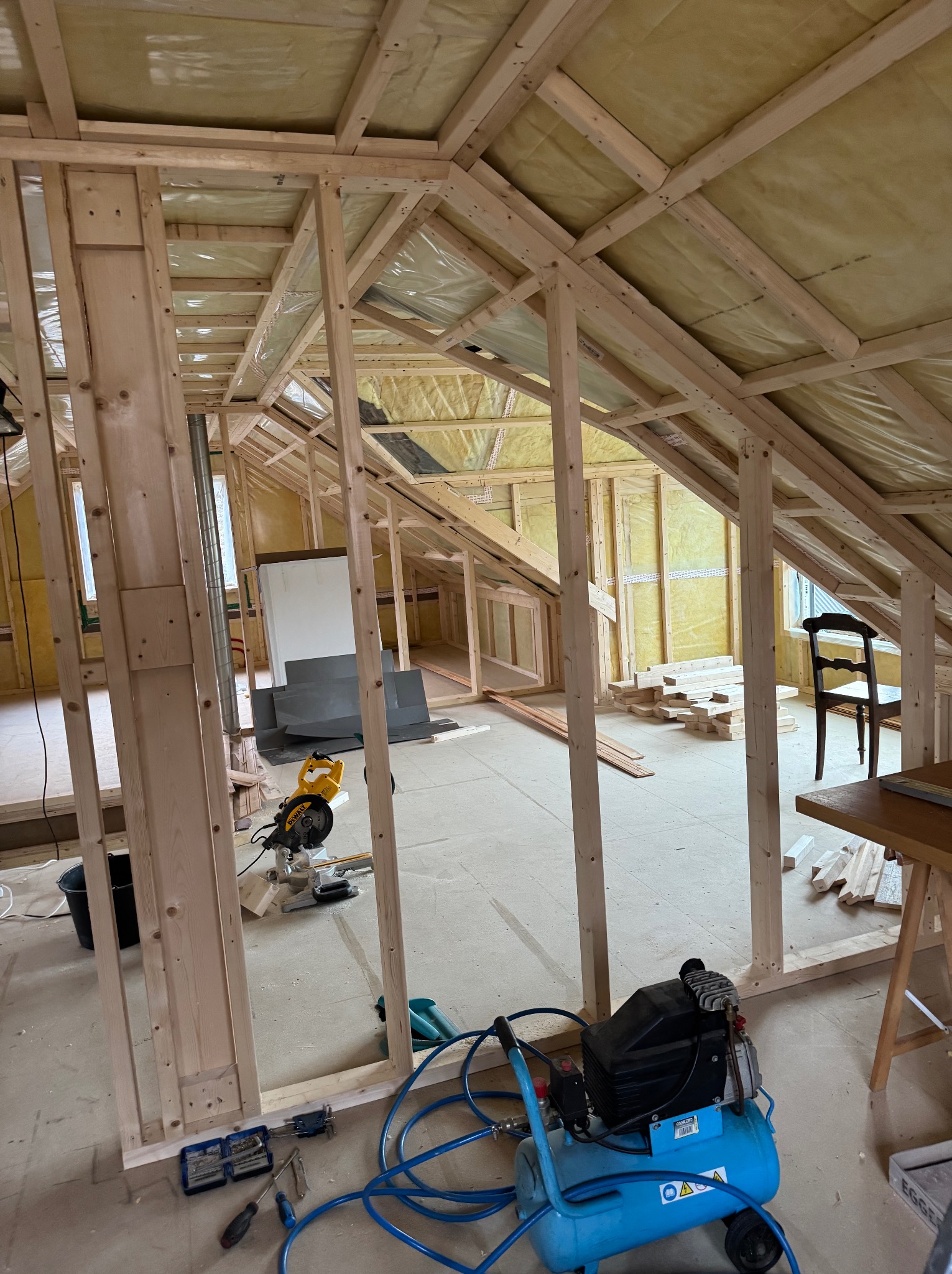 Wnętrze domu szkieletowego w trakcie budowy: drewniana konstrukcja, izolacja z wełny mineralnej, narzędzia budowlane, kompresor. Widok na poddasze z oknami.