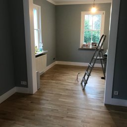 Krzysztof Poleszak - Jasny parkiet w pokoju z szarymi ścianami i białymi listwami. Widoczne okna, drabina i materiały budowlane. Delikatne światło dzienne.