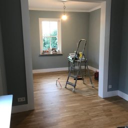 Krzysztof Poleszak - Pokój po remoncie z drewnianą podłogą, drabina i narzędzia. Widok przez białe obramowanie przejścia, okno w tle. Ściany w odcieniach szarości.