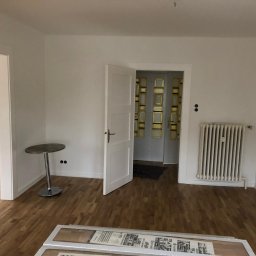 Krzysztof Poleszak - Jasne wnętrze po remoncie: parkiet, białe ściany, grzejnik, otwarte drzwi. Na podłodze panele w opakowaniu. Widok na sąsiednie pomieszczenia.