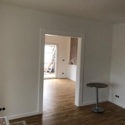 Krzysztof Poleszak - Jasne wnętrze po remoncie: świeżo pomalowane ściany, parkiet, widoczne gniazdka elektryczne. W tle drabina i okno balkonowe. Minimalistyczny stolik.