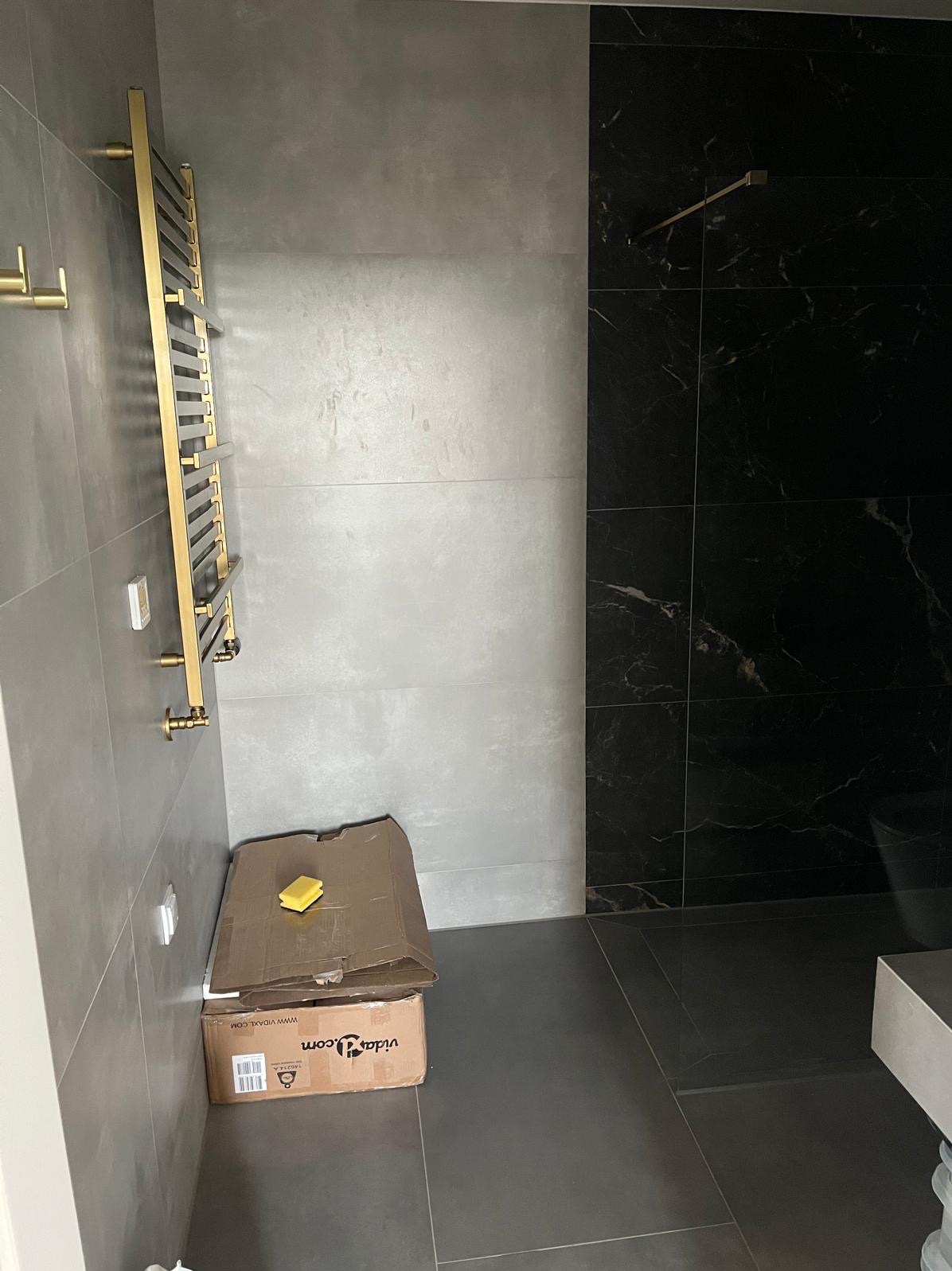 Nowoczesna łazienka z szarymi i czarnymi płytkami, złotym grzejnikiem drabinkowym i akcesoriami. Na podłodze karton z gąbką. Minimalistyczny design.