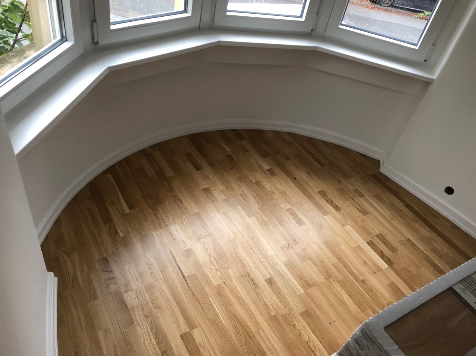 Jasny parkiet ułożony w pokoju z oknem wykuszowym. Widoczne listwy przypodłogowe i fragment opakowania paneli. Ujęcie z góry, naturalne światło.