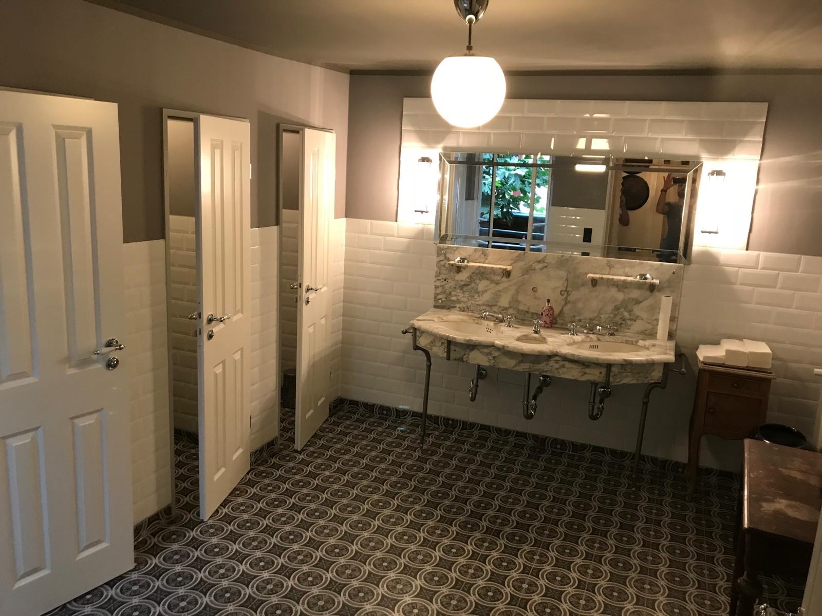 Wykończona łazienka w stylu retro: białe płytki, marmurowa umywalka z dwoma kranami, wzorzysta podłoga i białe drzwi do kabin. Oświetlenie punktowe i lustro.