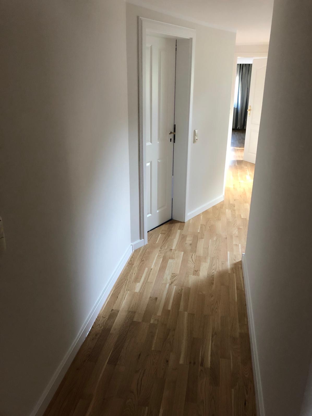 Jasny korytarz z parkietem ułożonym w jodełkę, białe ściany i drzwi. Widok w głąb mieszkania z otwartymi drzwiami i zasłonami w tle.