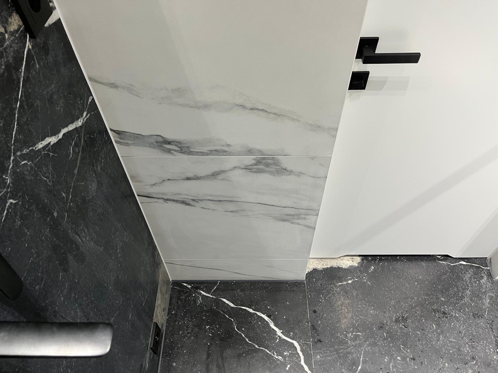 Fragment wykończonej łazienki z czarno-białymi płytkami imitującymi marmur na ścianach i podłodze, widoczny czarny uchwyt drzwiowy.