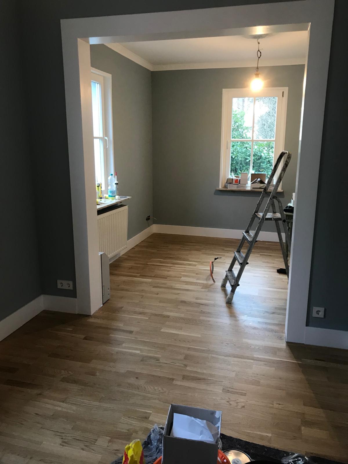 Jasny parkiet w pokoju z szarymi ścianami i białymi listwami. Widoczne okna, drabina i materiały budowlane. Delikatne światło dzienne.