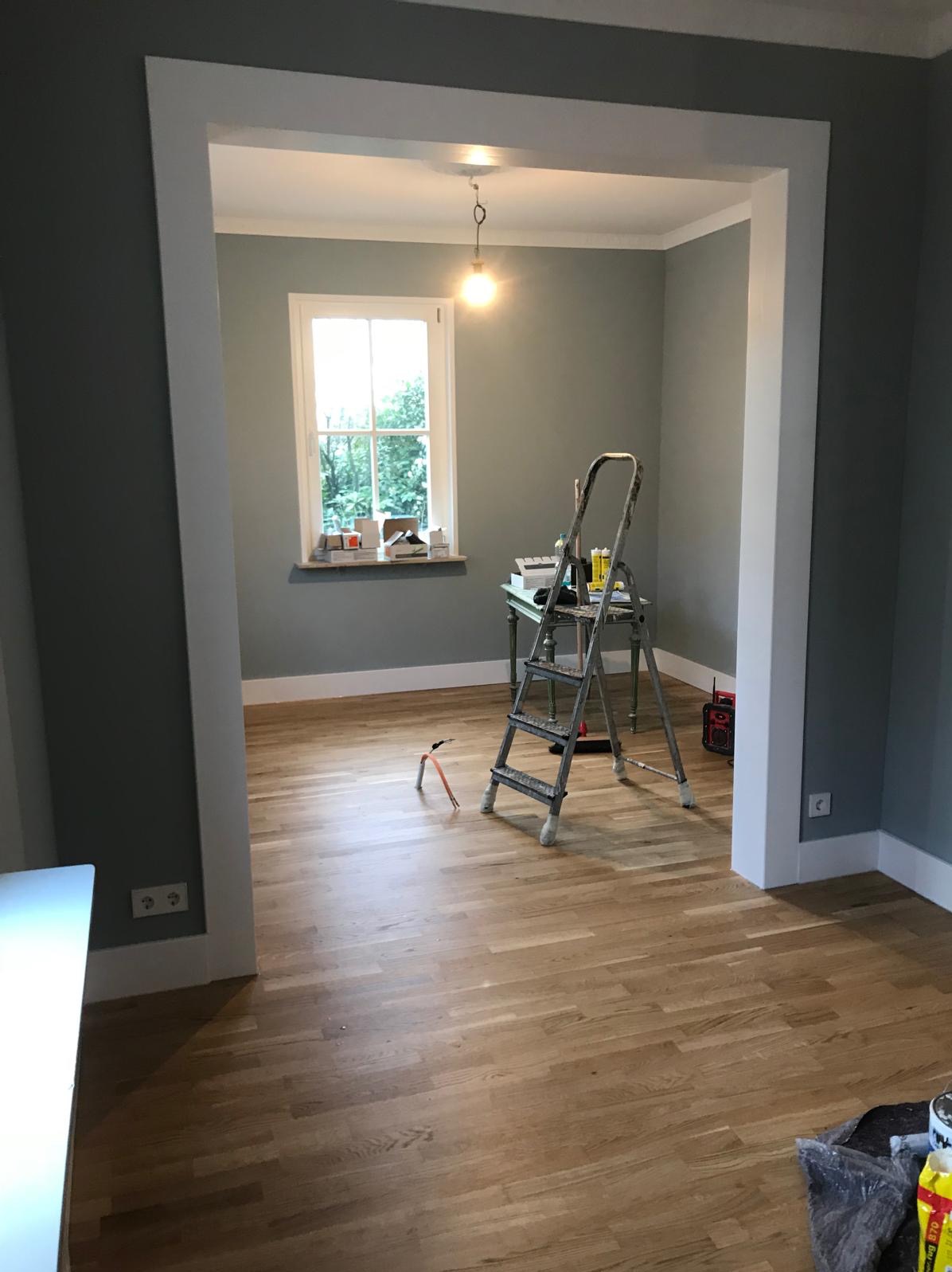Pokój po remoncie z drewnianą podłogą, drabina i narzędzia. Widok przez białe obramowanie przejścia, okno w tle. Ściany w odcieniach szarości.