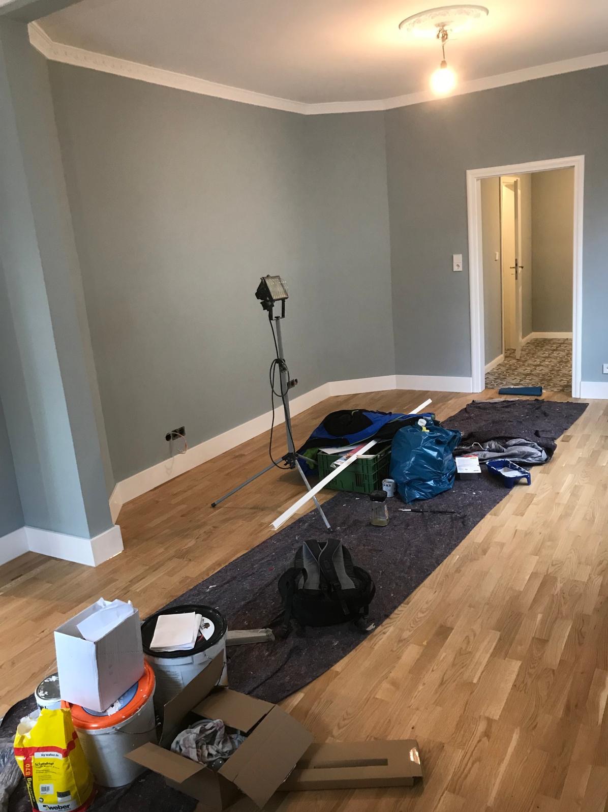 Pokój w trakcie malowania ścian na szaro, z widocznymi narzędziami malarskimi, lampą na statywie i zabezpieczonym parkietem. Listwy przypodłogowe i sufitowe w kolorze białym.