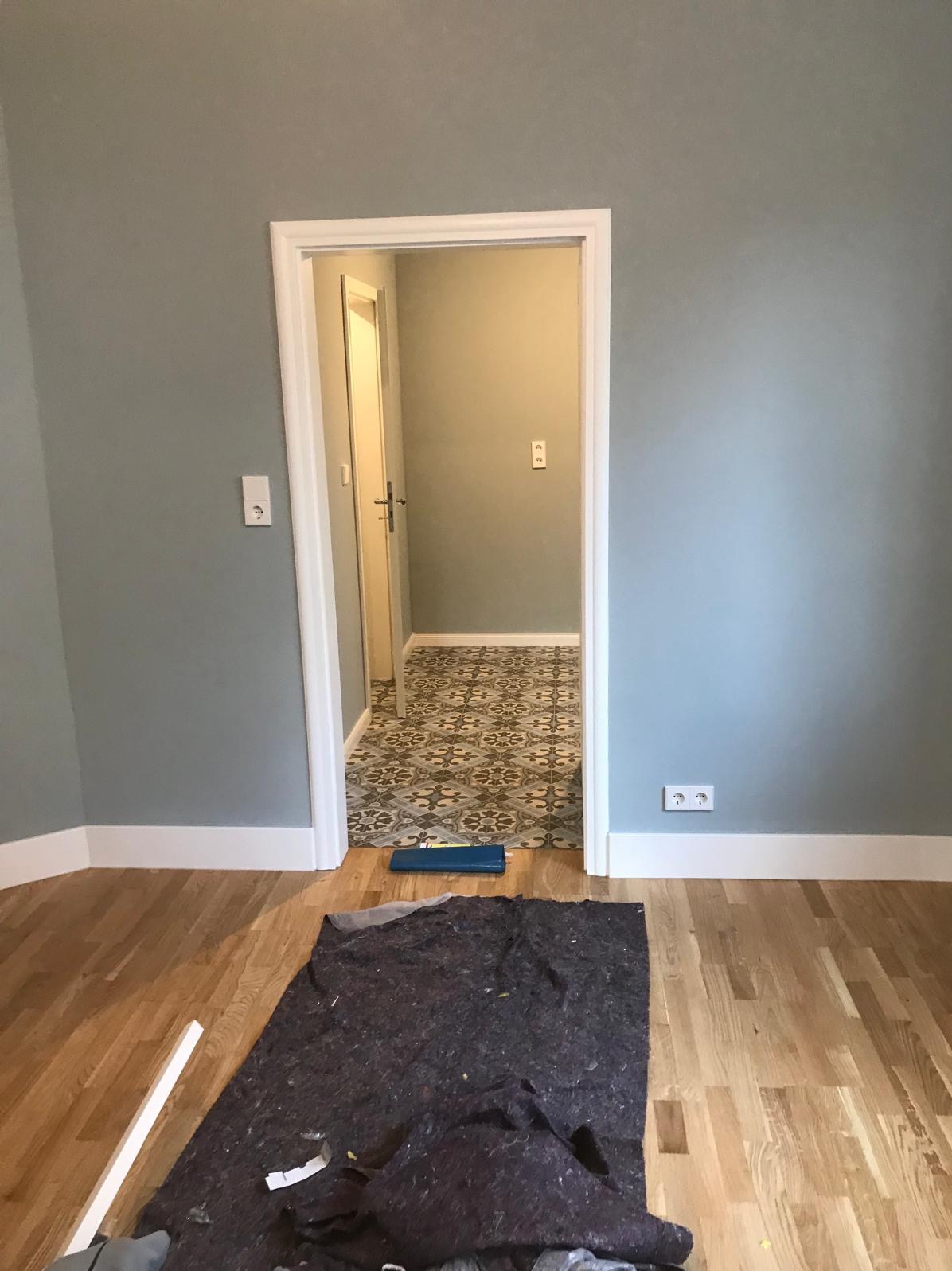 Pokój z drewnianą podłogą i widokiem na korytarz z wzorzystą posadzką przez białe drzwi. Ochronna mata na podłodze, ściany w odcieniach szarości.