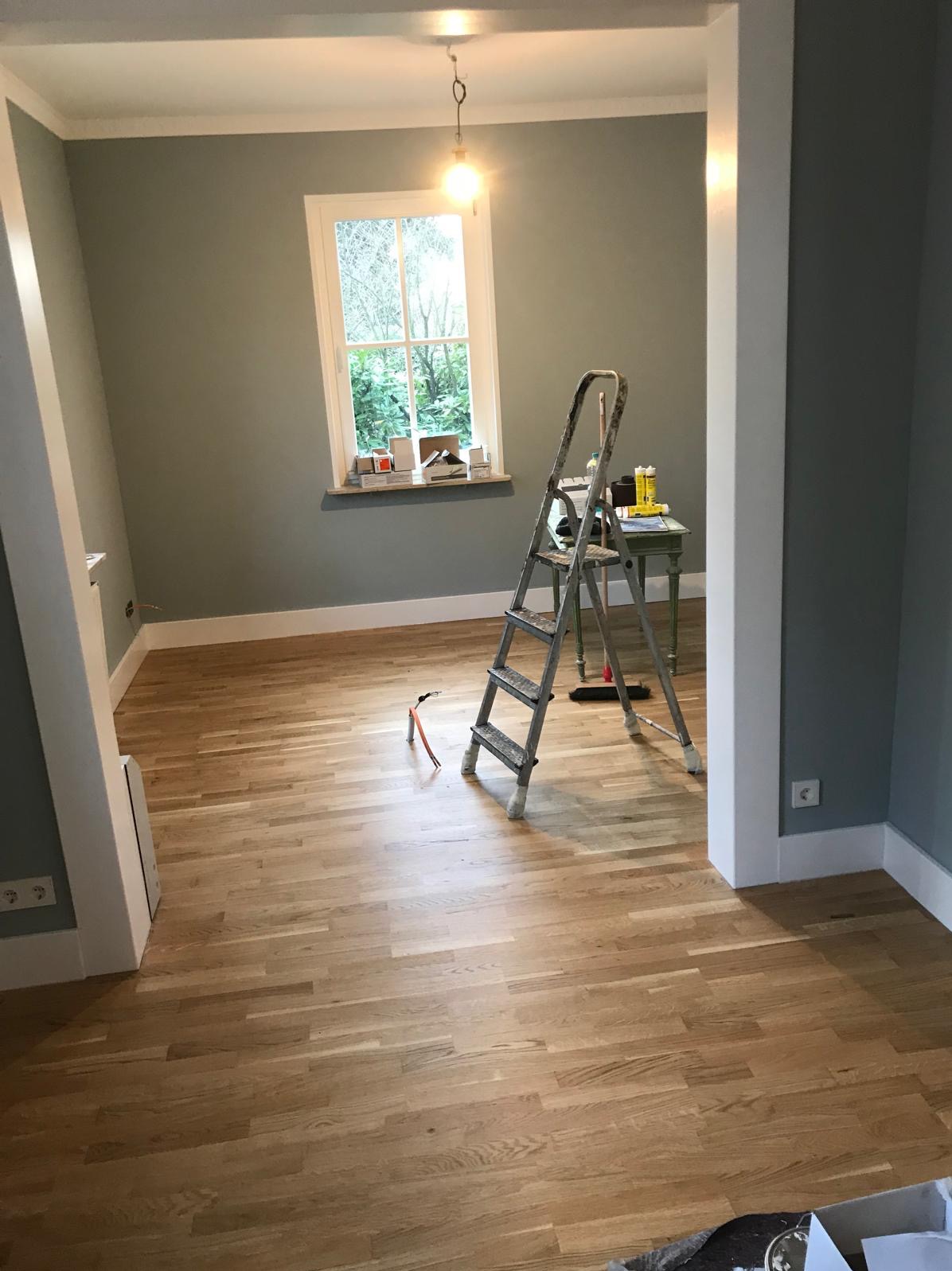 Jasny parkiet w pokoju z szarozielonymi ścianami, drabina i narzędzia. Widok na okno z zielenią. Listwy przypodłogowe i sufitowe w kolorze białym. Gniazdka elektryczne.