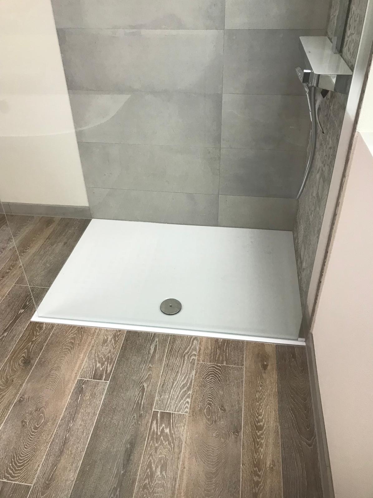 Nowoczesna kabina prysznicowa z brodzikiem i szarymi płytkami ściennymi. Podłoga wyłożona panelami imitującymi drewno. Minimalistyczny design.