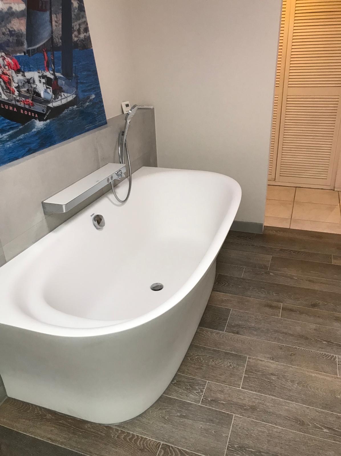 Nowoczesna, wolnostojąca wanna w jasnej łazience z drewnopodobną podłogą i obrazem żaglówki. Minimalistyczny design, chromowana armatura, subtelne oświetlenie.