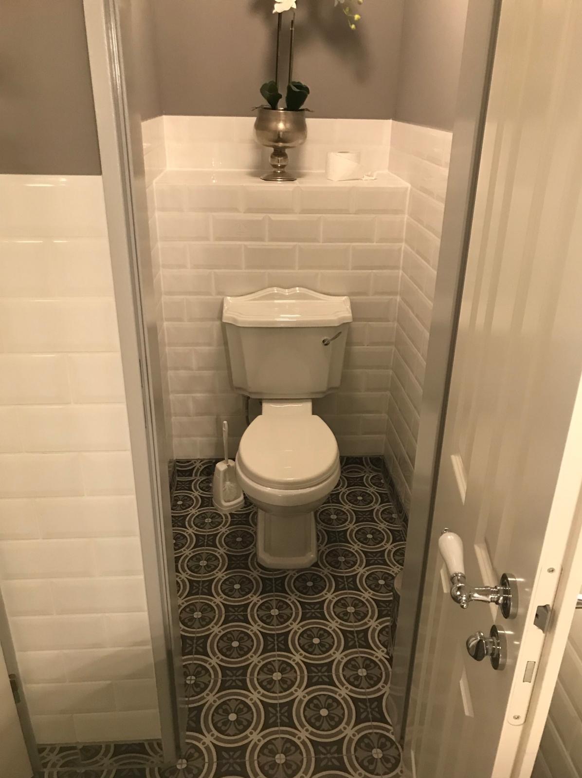 Kompaktowa toaleta z białą ceramiką, mozaikową podłogą i subtelną dekoracją kwiatową. Ściany wyłożone białymi, prostokątnymi płytkami.