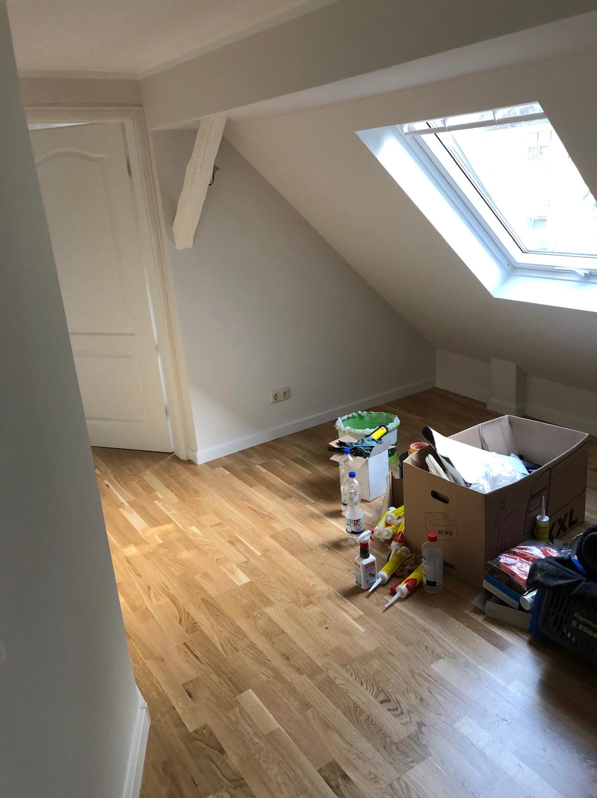 Poddasze z oknem dachowym i świeżo ułożonym parkietem. Widoczne materiały budowlane i narzędzia, sugerujące prace wykończeniowe w toku. Jasne drewno parkietu.