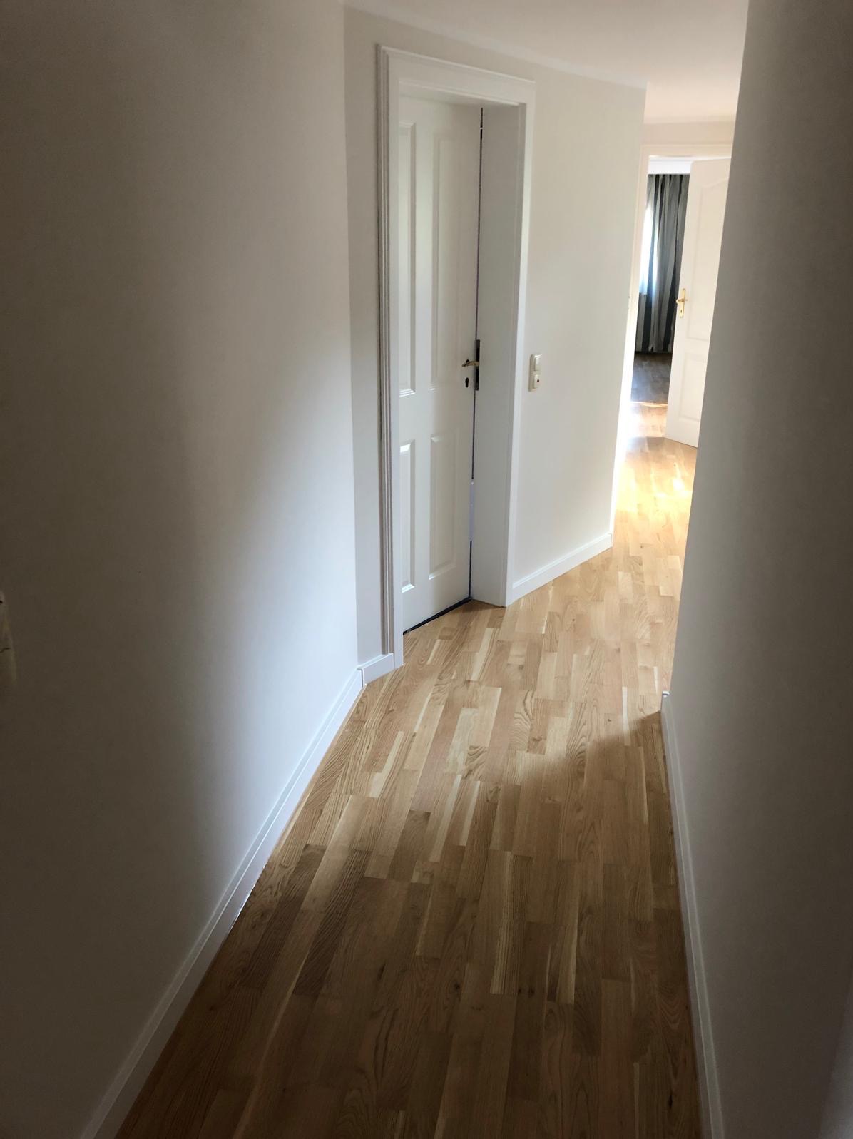 Jasny korytarz po remoncie: parkiet z naturalnego drewna, białe ściany i drzwi. Widok w głąb mieszkania z zasłonami w tle. Minimalistyczny design.