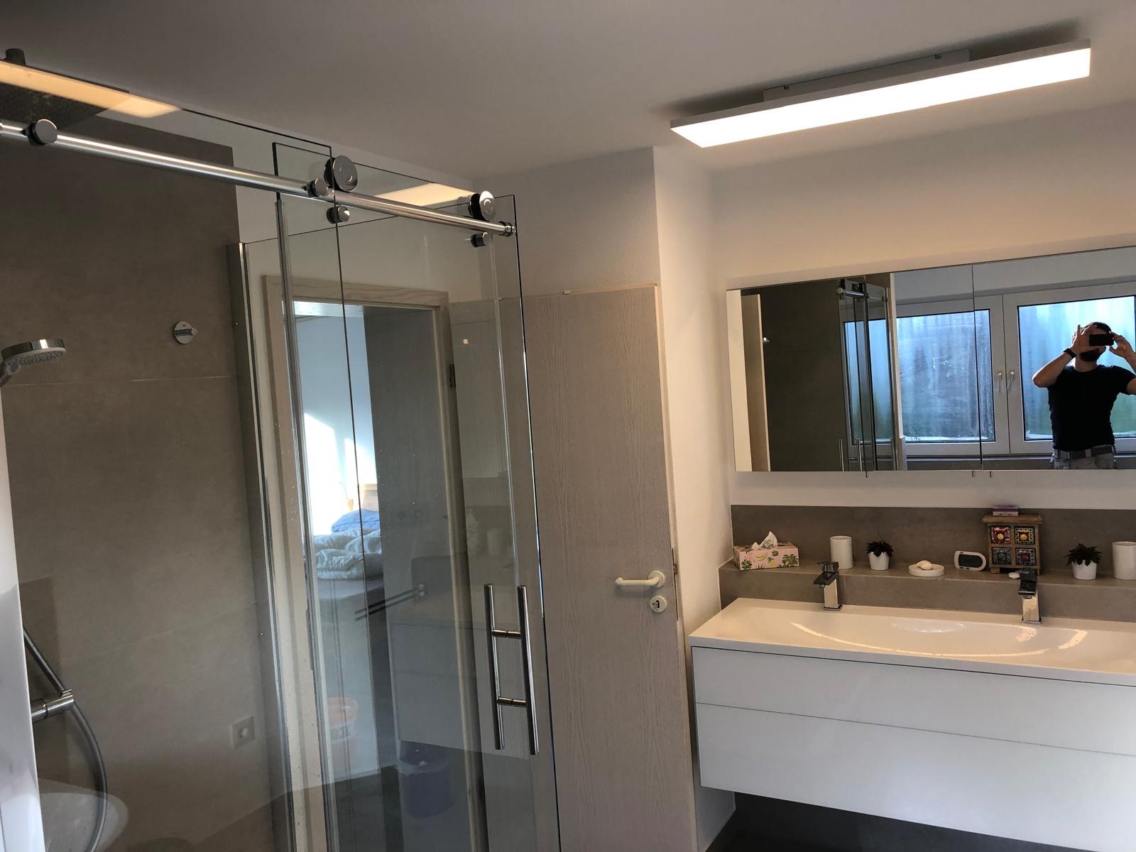 Nowoczesna łazienka z kabiną prysznicową typu walk-in, umywalką z szafką i dużym lustrem z oświetleniem. Widoczne szare płytki i minimalistyczne wykończenie.