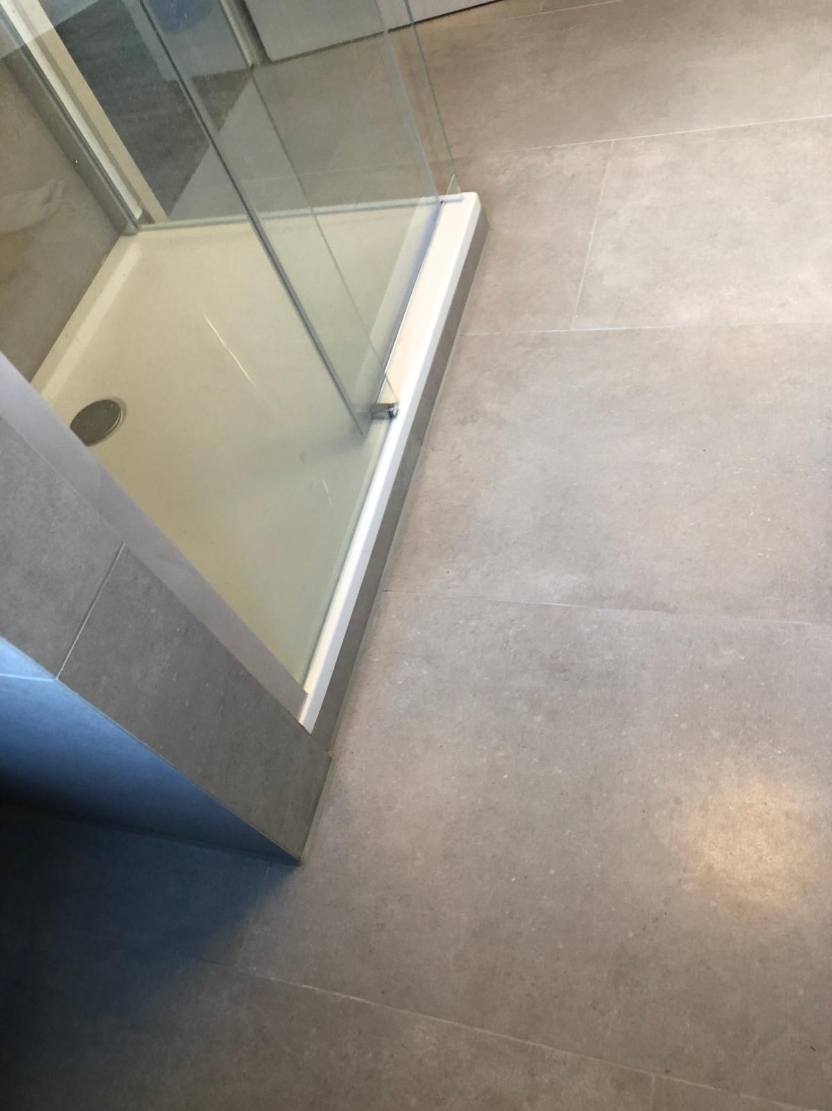 Szara, matowa podłoga z płytek w łazience, z widoczną kabiną prysznicową z białym brodzikiem i szklanymi ściankami. Minimalistyczny design, stonowana kolorystyka.