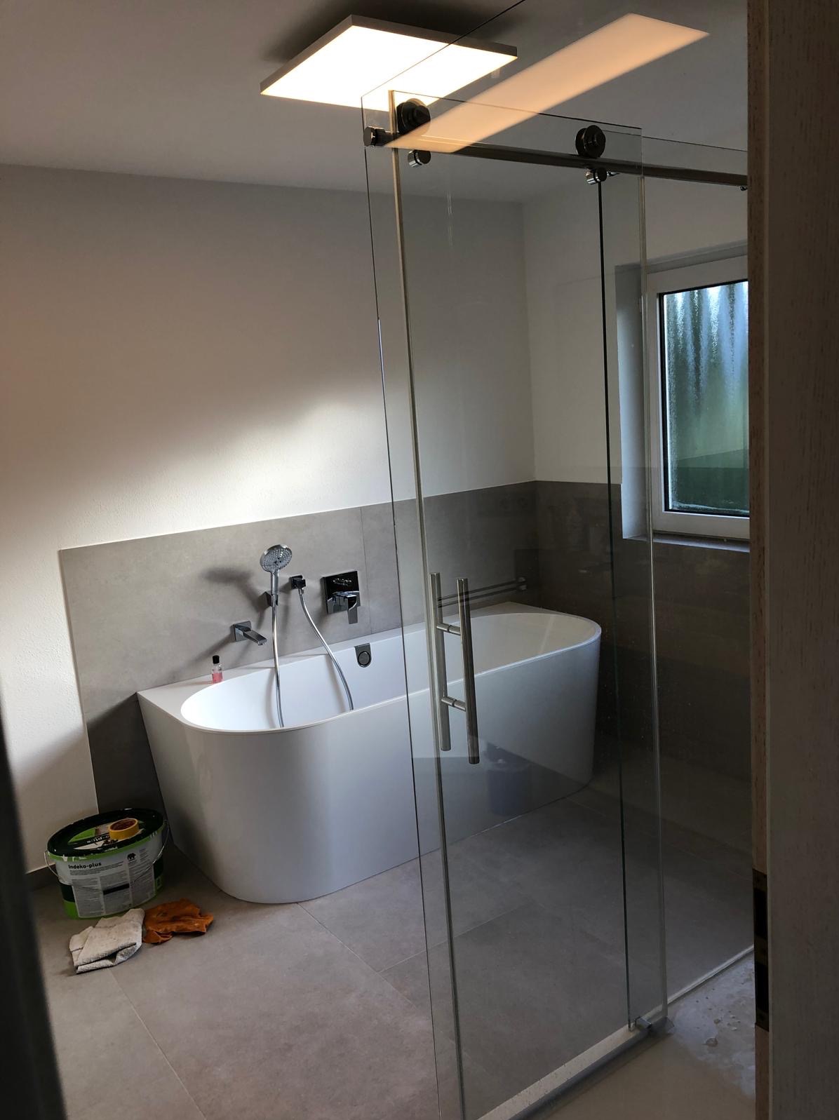 Nowoczesna łazienka z wolnostojącą wanną i szklaną kabiną przesuwną. Widoczny fragment ściany z szarą okładziną i bateria wannowa. Na podłodze płytki i wiadro z farbą.