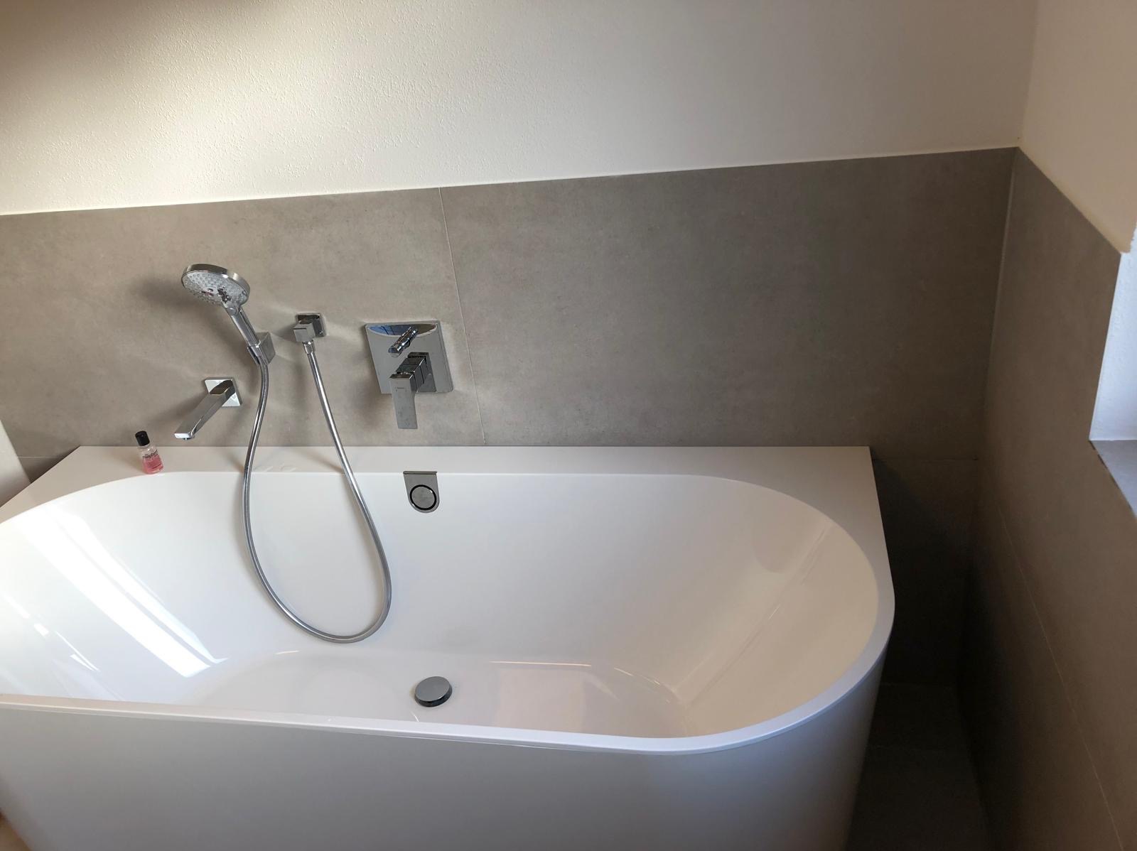 Nowoczesna, biała wanna z chromowaną armaturą i szarymi płytkami w tle. Minimalistyczny design, czyste linie, mała buteleczka kosmetyku na rancie wanny.