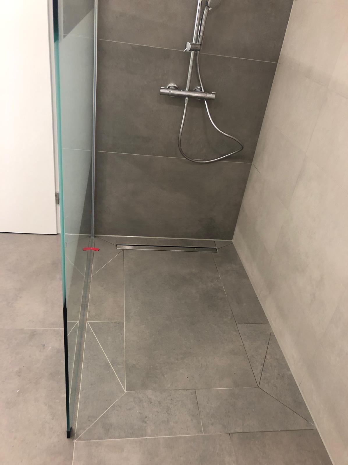 Nowoczesna kabina prysznicowa z szarymi płytkami, odpływem liniowym i szklaną ścianką. Widoczne detale wykończenia i precyzyjne ułożenie płytek pod kątem.