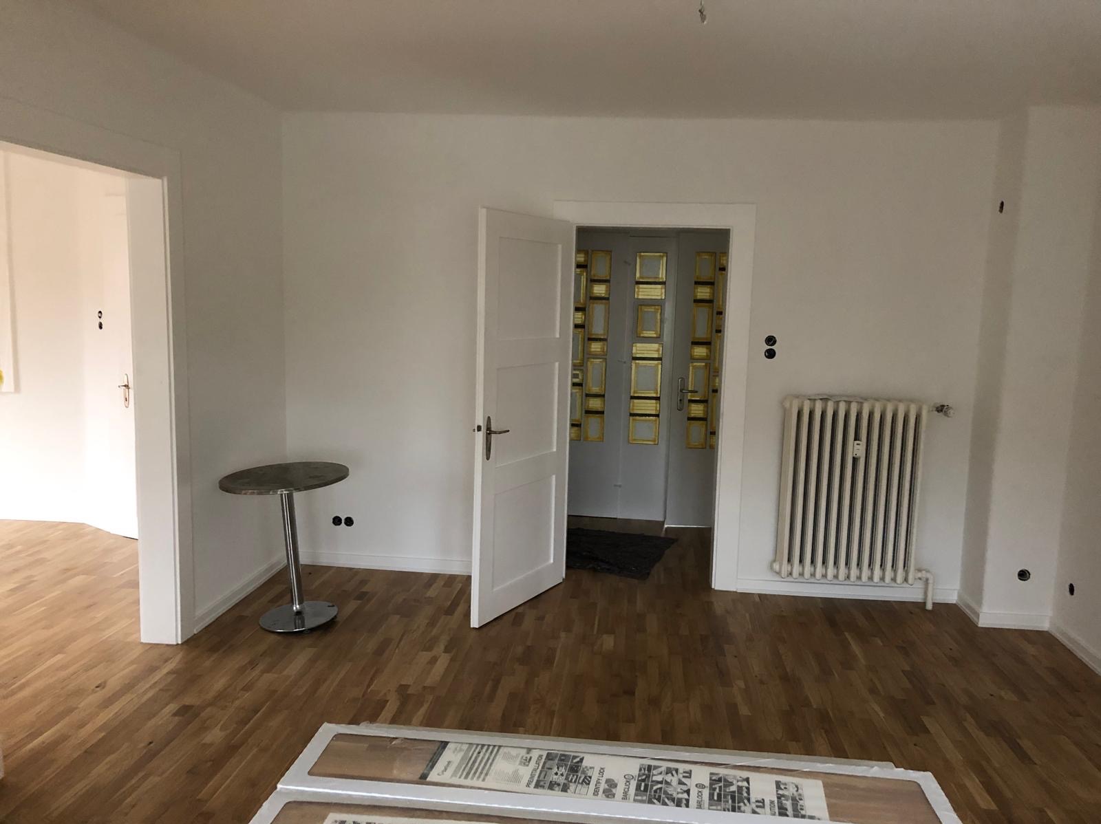 Jasne wnętrze po remoncie: parkiet, białe ściany, grzejnik, otwarte drzwi. Na podłodze panele w opakowaniu. Widok na sąsiednie pomieszczenia.