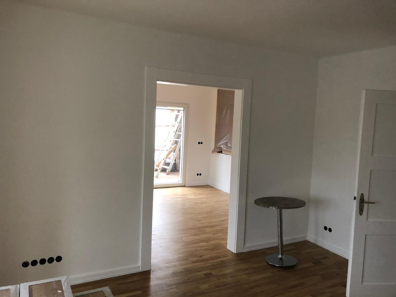 Jasne wnętrze po remoncie: świeżo pomalowane ściany, parkiet, widoczne gniazdka elektryczne. W tle drabina i okno balkonowe. Minimalistyczny stolik.