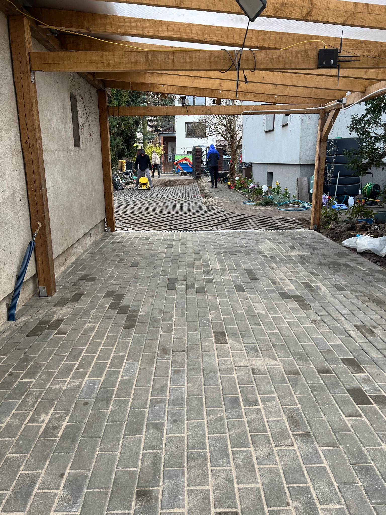 Ułożona szara kostka brukowa na podjeździe pod wiatą, w tle pracownik zagęszczarką wibruje nawierzchnię z ażurowych płyt. Widoczne domy i zieleń.