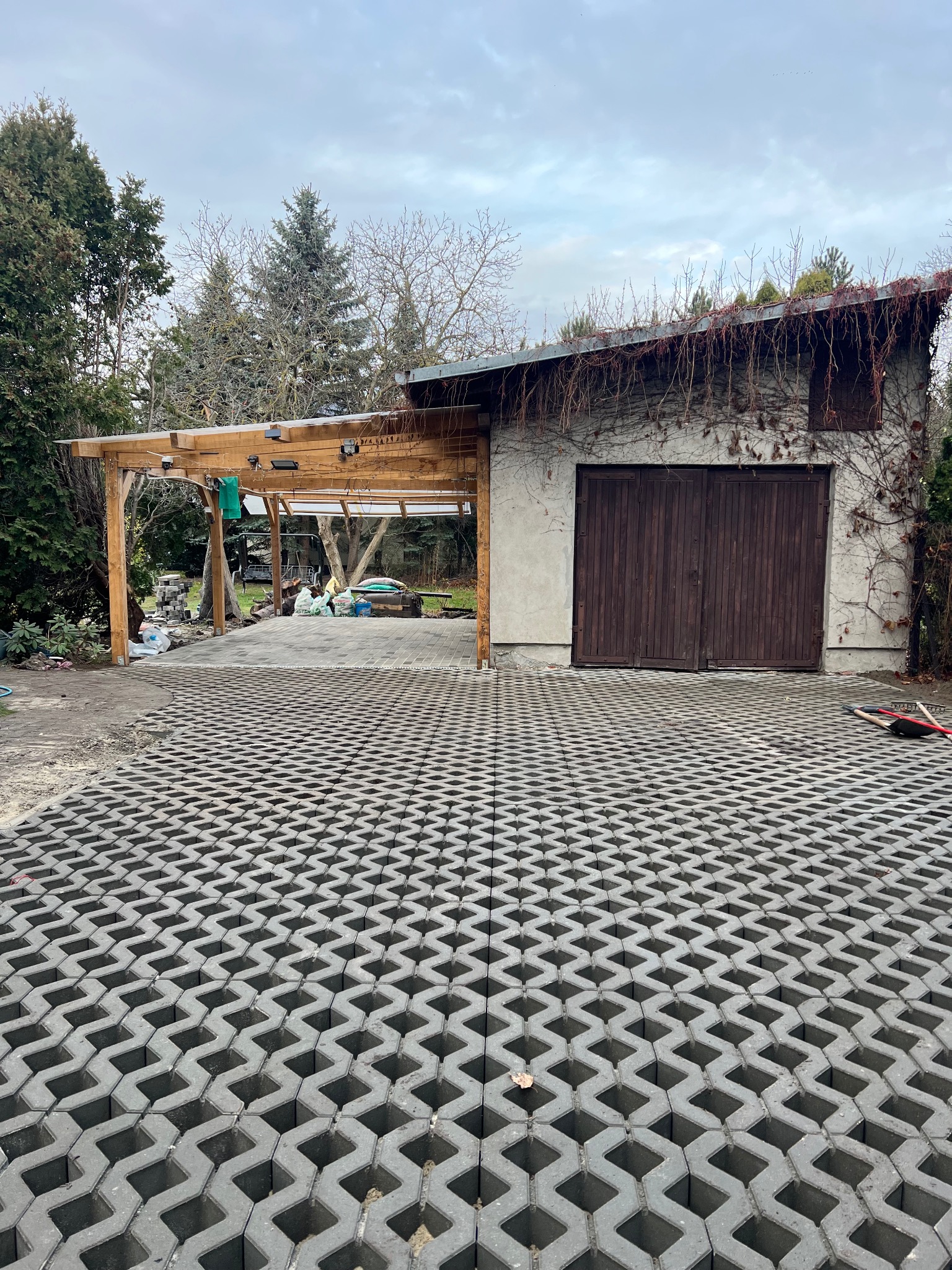 Ułożona ażurowa kostka brukowa przed garażem i drewnianą wiatą. Widoczne narzędzia i materiały budowlane. Pochmurne niebo w tle.