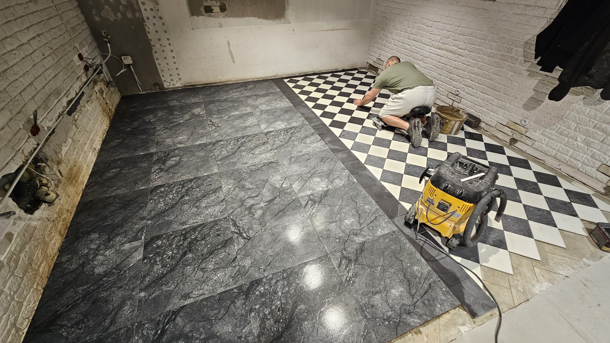 Układanie podłogi z płytek: szare marmurowe i czarno-biała szachownica. Glazurnik na kolanach, odkurzacz budowlany obok. Częściowo wykończone pomieszczenie.