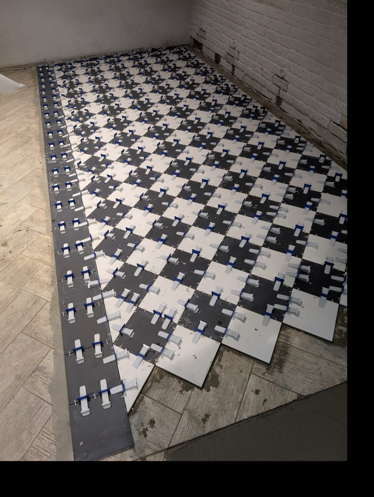 Układanie podłogi w szachownicę z płytek czarno-białych, z użyciem systemu poziomowania płytek z niebieskimi klinami. Widoczny fragment ściany z cegły.