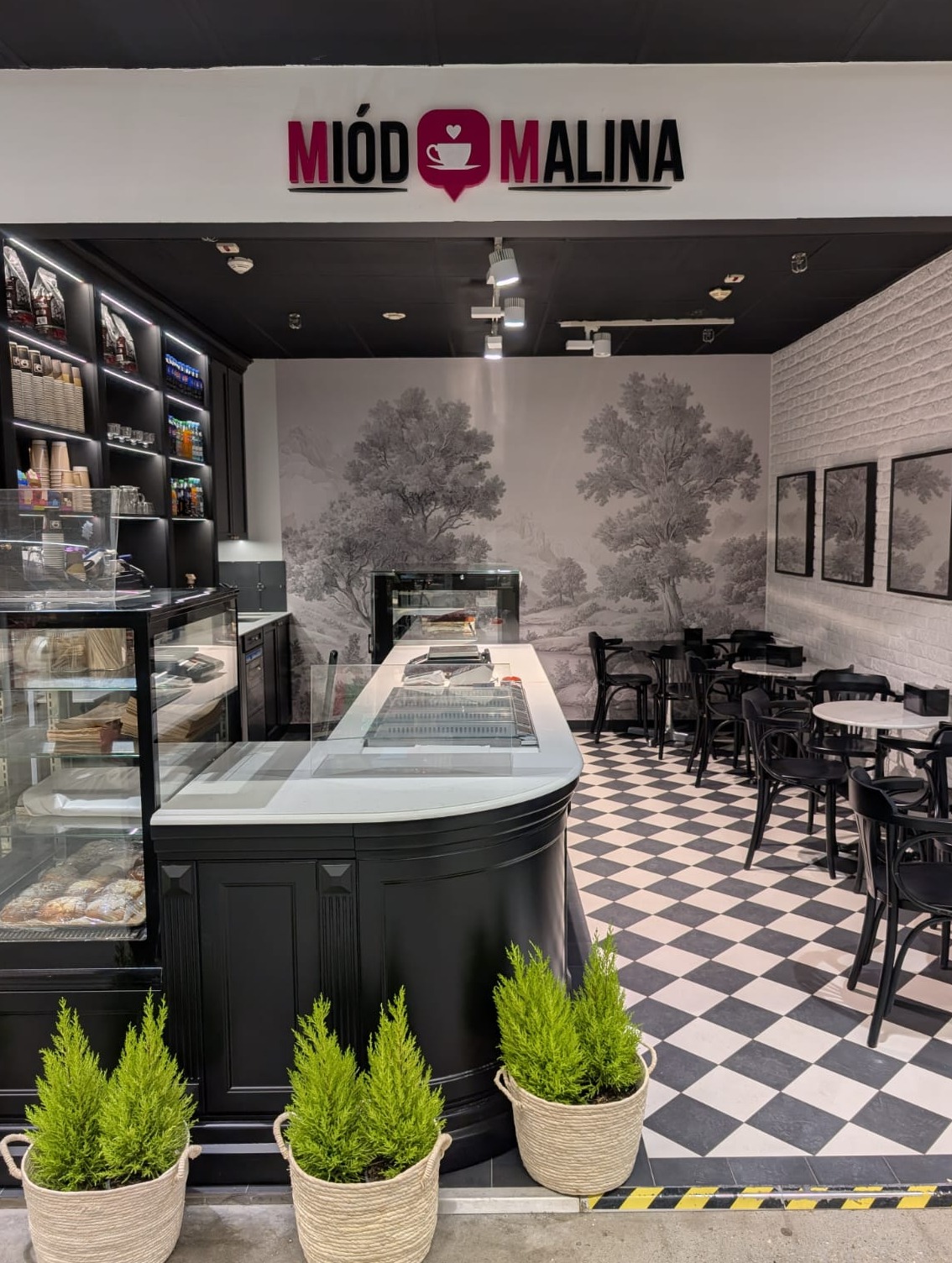 Eleganckie wnętrze kawiarni z czarno-białą szachownicą na podłodze, stylowym kontuarem i dekoracyjną tapetą z motywem drzew. Widoczne stoliki i krzesła.
