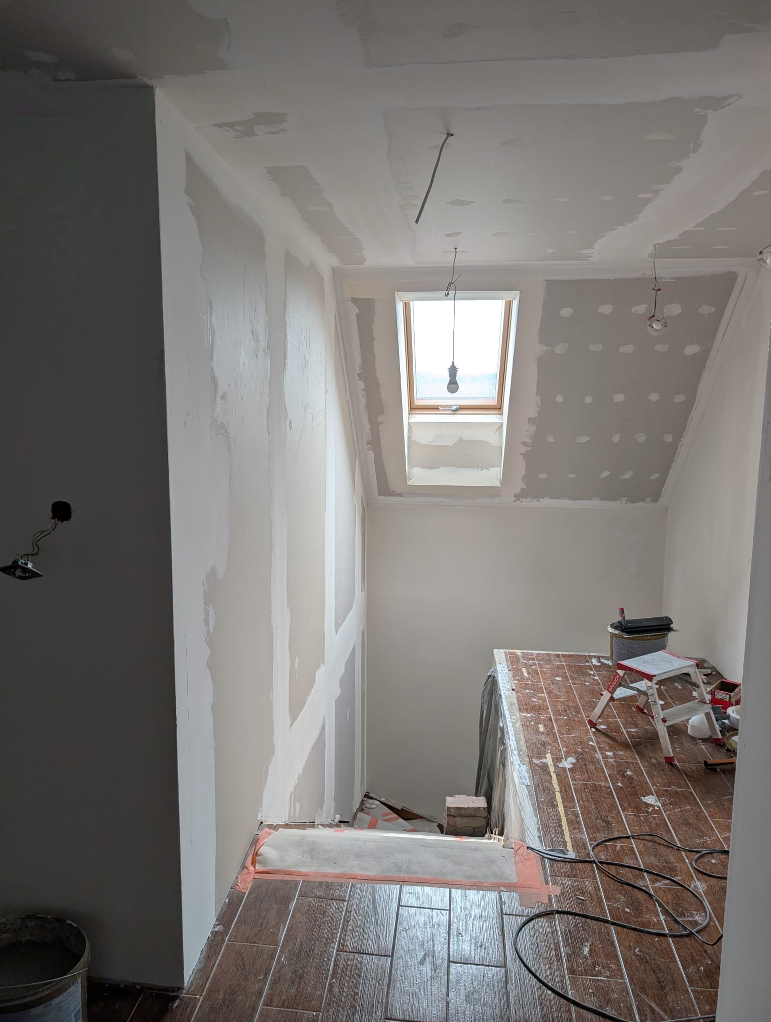 Poddasze w trakcie remontu: szpachlowane ściany, okno dachowe, zabezpieczona podłoga z płytek imitujących drewno, drabina malarska i narzędzia. Widok z góry.