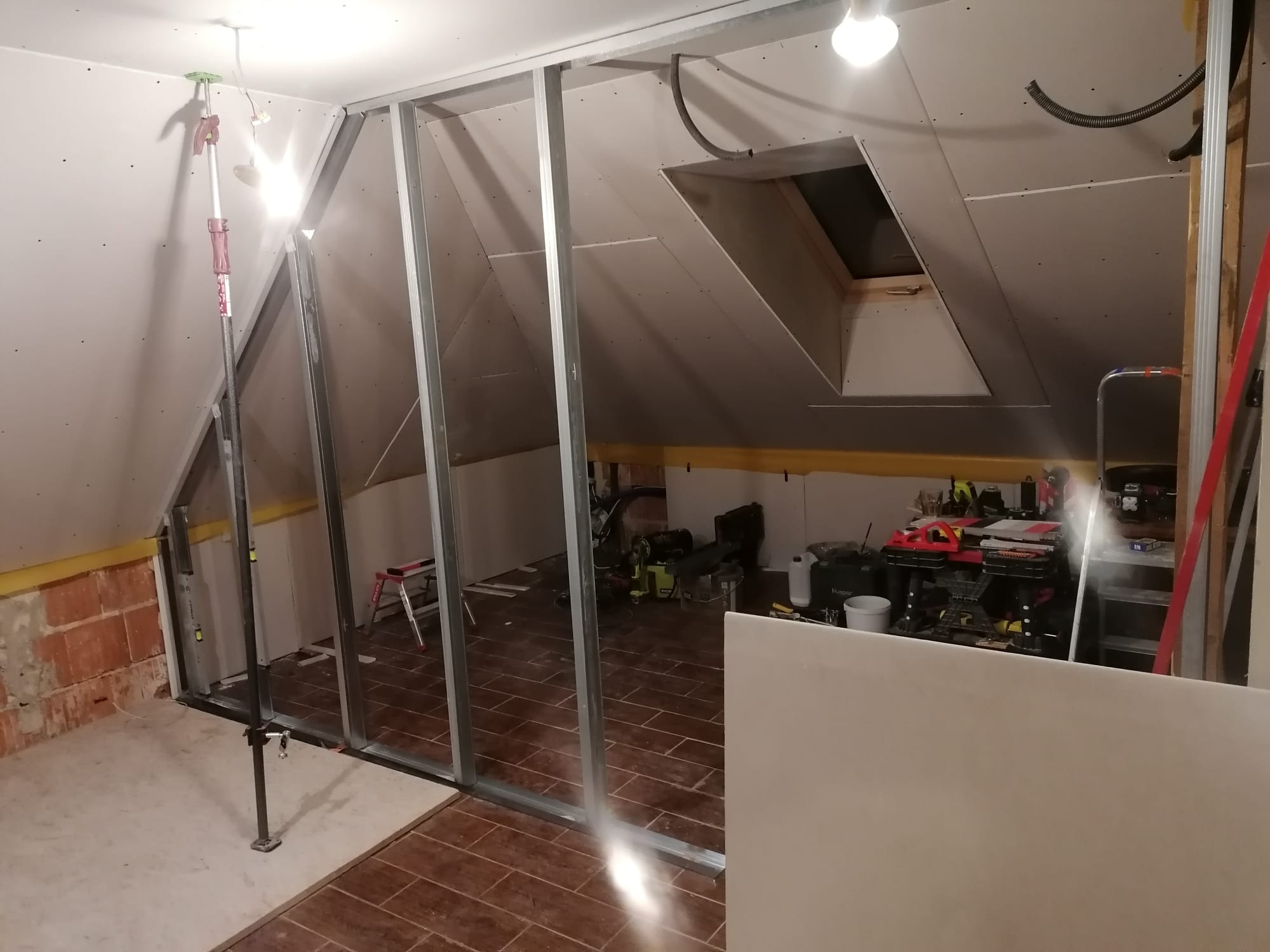 Montaż ścianki działowej z płyt gipsowo-kartonowych na poddaszu. Widoczne metalowe profile i częściowo zamontowane płyty. W tle narzędzia i okno dachowe.