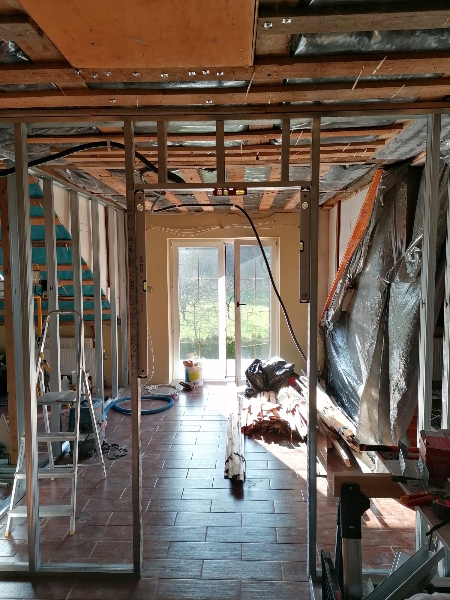 Konstrukcja szkieletowa z profili stalowych w trakcie montażu, widoczne poziomice, narzędzia i materiały budowlane. Podłoga wyłożona płytkami imitującymi drewno.