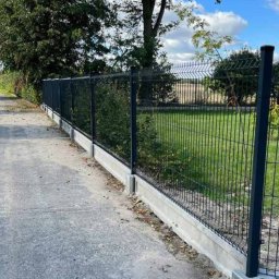 Ogrodzenia Systemowe - Ciemna, panelowa konstrukcja ogrodzeniowa z podmurówką betonową, widoczna z boku na tle zieleni i błękitnego nieba. Słupki ogrodzenia w kolorze grafitowym.
