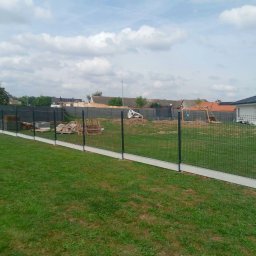 Ogrodzenia Systemowe - Nowoczesne ogrodzenie panelowe z podmurówką betonową na zielonym trawniku. W tle domy jednorodzinne i błękitne niebo z chmurami. Widok z poziomu gruntu.