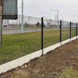 Ogrodzenia Systemowe - Długi, panelowy płot z podmurówką betonową, ciągnący się wzdłuż drogi. Słupki i panele w kolorze grafitowym. Trawa i ziemia na pierwszym planie, budynki w tle.