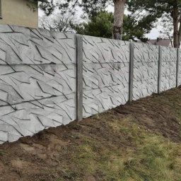 Ogrodzenia Systemowe - Długi, szary mur z betonu z geometrycznym wzorem, oddzielający posesję od drogi. Widoczne słupki i panele. Trawa i ziemia u podnóża. W tle dom i drzewa.