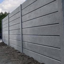 Ogrodzenia Systemowe - Szary, prefabrykowany mur z betonu architektonicznego, z poziomymi żłobieniami, tworzący nowoczesne ogrodzenie posesji. Widok z dołu, z ziemią u podstawy.